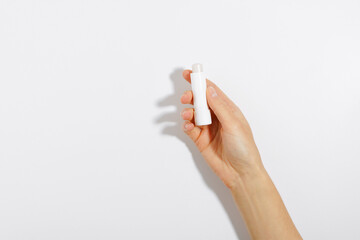 Female hand holding white moisturizing lip balm on white isolated background with shadow from direct light. The concept of beauty products and decorative cosmetics for the face - obrazy, fototapety, plakaty