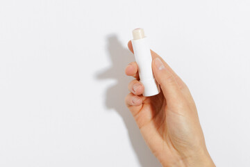 Close-up of a woman’s hand holding a transparent mockup lip balm in a white package on a white isolated background. Concept of skin care, beauty and health products, sun and cold protection - obrazy, fototapety, plakaty