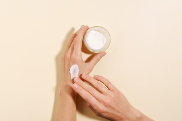 Close-up of a female hand holding a glass jar of moisturizer and applying a smear on the skin on a beige isolated background. Concept of beauty products, face care