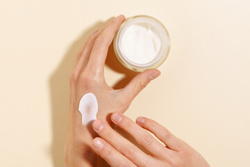 Female fragile hands holding a glass jar of facial cream and smearing it on the skin on a beige...