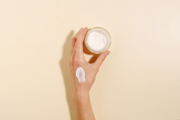 Female hand with cream smear holding glass jar of moisturizer on beige isolated background. The...