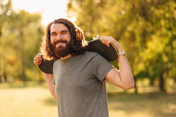 Photo of young bearded hipster man holding skateboard and looking at the camera in park during...
