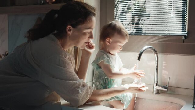 Young Mother and Little Baby Child Playing with Water Flowing from a Tap