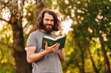 Photo of young bearded hipster man with long hair reading a book in park in sunset light.