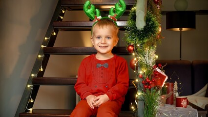 Happy cute toddler boy in pajamas and antlers smiling in camera and having fun on Christmas eve at house decorated for New Year. Family celebrations on winter holidays.
