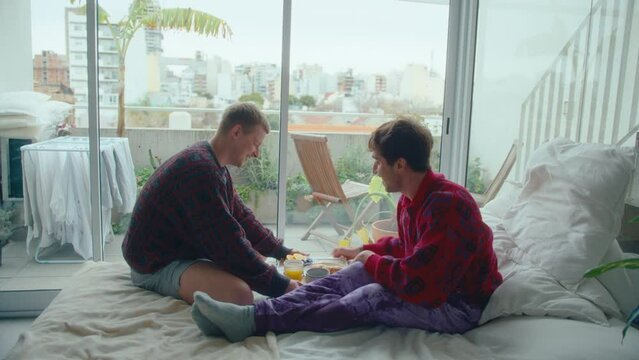 Young gay man bringing breakfast in bed for his happy boyfriend. Couple talking, kissing and enjoying meal in the morning at home. Zoom shot, handheld camera
