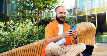 Portrait of handsome stylish young man in orange shirt and glasses and headphones using smartphone,...