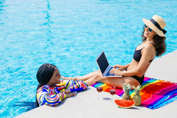 Business working woman with friend relax at luxury hotel resort swimming pool using laptop computer remote wireless working while summer holiday vacation. Freelance bikini female works at poolside.
