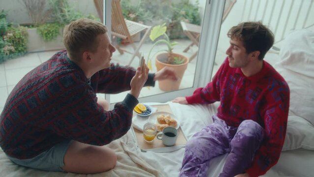 Young gay man feeding his boyfriend with blueberries and taking photos with phone while enjoying breakfast in bed at home. High angle view, handheld camera shot