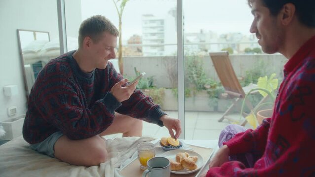 Young romantic gay man sitting on bed, feeding his boyfriend with fresh berries and taking pictures with smartphone while having fun during breakfast at home