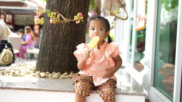Little asian Thai girl enjoying eat ice cream wear raditional Thai suit in buddhist temple outdoor summer vacation