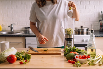 Lady in the kitchen presses a lime into a blender on a wooden table on which there are fresh vegetables.