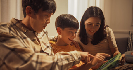 Beautiful Family of Three Using Digital Tablet Computer while Sitting on the Sofa. Father, Mother and Cute Little Boy Play Educational Video Games Together. Close Up Shot in the Evening.