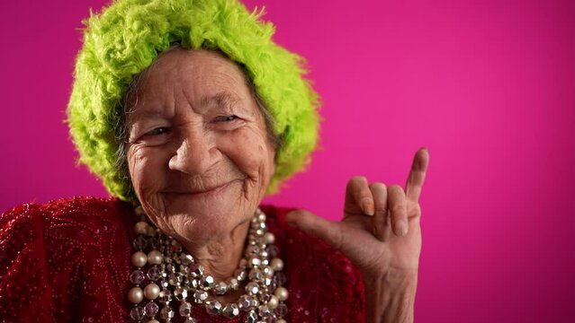 Fisheye view of funny elderly mature woman, 80s, wearing green wig giving SHAKA hand gesture isolated on pink background. Concept of old rock and roll person in slow motion