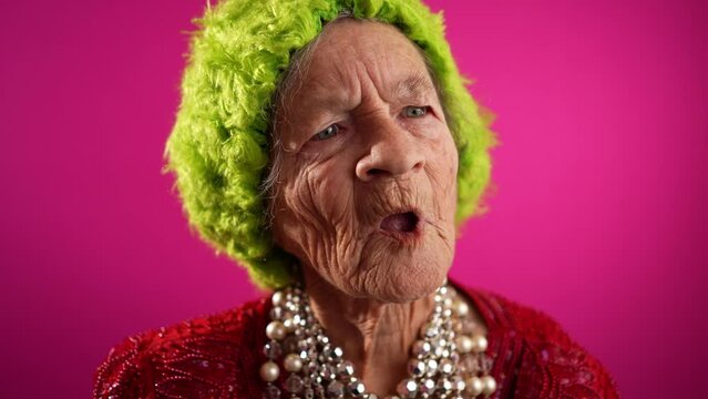 Slow motion funny view of mature elderly woman, 80s, having giving OKAY gesture, wearing green wig or hat isolated on pink background.