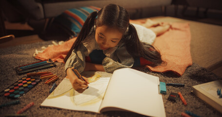 Evening Portrait of Cute Little Girl Drawing while Lying on the Floor in Living Room. Talented Korean Child Being Creative, Coloring Picture, Preparing to Become Famous Artist