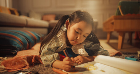Evening Portrait of Cute Little Girl Drawing while Lying on the Floor in Living Room. Talented...