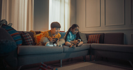 Cute Little Siblings Playing Video Game On Gaming Station On The Sofa In Cozy Living Room. Korean Brother And Sister Enjoying Games Together In The Evening. Children Having Fun At Home After School. - obrazy, fototapety, plakaty