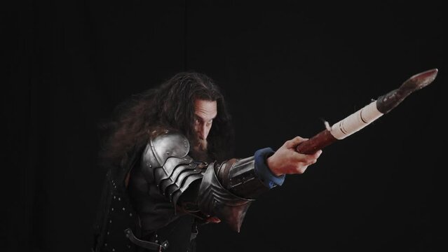 A charismatic man in the armor of a medieval knight hits with a spear on a black background.