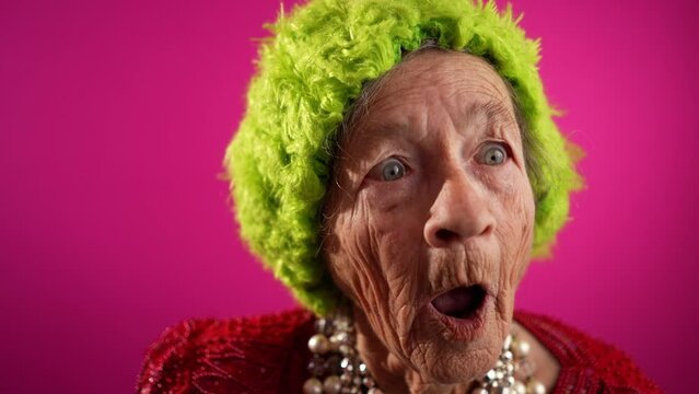 Saying WOW, a happy fisheye caricature of funny elderly woman with green hat isolated on pink background in slow motion