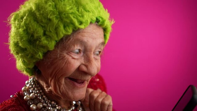 Funny fisheye view of old woman with no teeth having video chat on phone with family and friends wearing green wig isolated on pink background in slow motion