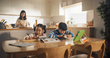 Portrait of a Little Korean Kids Sitting at a Kitchen Table at Home in the Morning While Their Mother is Cooking in the Background. Caring Brother Helping his Little Sister with her Homework
