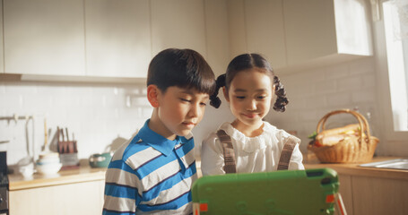 Portrait of Two Korean Kids Using a Digital Tablet During Weekend in The Kitchen. Two Cute Asian...