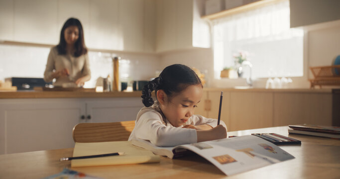 Portrait of a Korean Female Child Sitting at a Kitchen Table and Drawing While her Mother is Preparing Breakfast. Little Cute Girl Waiting for Nutritious Meal Before Going to School