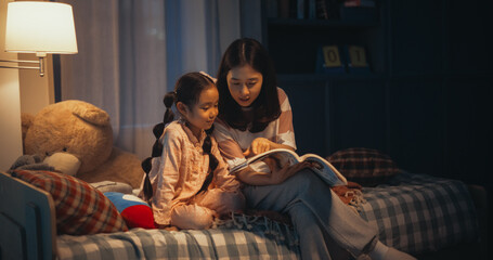 Korean Mother Reading a Fairytale to Her Lovely Small Daughter in Bed Before Going to Sleep. Young Beautiful Mom Caring for Little Girl, Reading Stories from a Book in the Evening at Home - obrazy, fototapety, plakaty