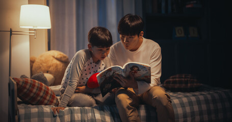 Korean Father Reading a Fairytale to His Lovely Cute Son in Bed Before Going to Sleep. Patient Father Helping his Son to Learn how to Read his Favorite Book. Parent and Child Bonding Moment - obrazy, fototapety, plakaty