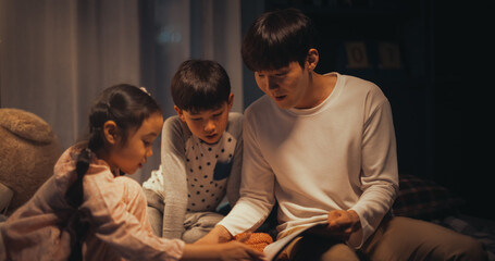 Korean Father Reading a Fairytale to His Lovely Cute Kids in Bed Before Going to Sleep. Young Handsome Dad Caring for his Little Children, Reading Stories from a Book in the Evening at Home