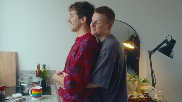 Young affectionate gay man hugging beloved boyfriend from behind and kissing him in the kitchen at home