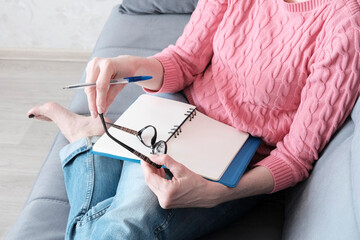 A woman in a pink sweater sits, holds glasses and a notepad with a pen for notes. Concept of online...