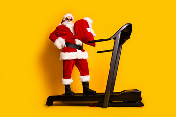 Full length photo of joyful funky man santa claus on sportive equipment holding full sack gifts isolated yellow color background
