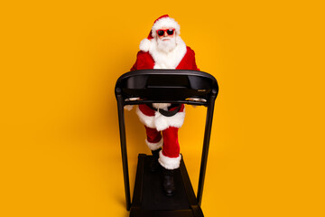 Full length photo of nice elderly man wear santa claus clothes running on treadmill gym isolated on yellow color background