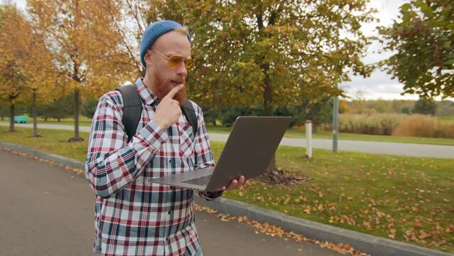 Young red-bearded man in yellow sun glasses, blue bini putting point finger to his mouth, touching his beard being in deep thoughts, typing on laptop. High quality 4k footage