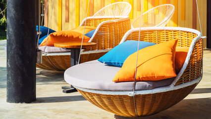 Colourful pillows on modern daybed. Brown rattan loungers with orange and blue pillows hanging with rope by relaxing terrace. Sunbed for sunbathing and swimming pool. Beautiful circular wooden daybed.