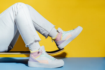 Close up female legs in white jeans and retro style high-top multicolor sport sneakers shoes sitting on the skateboard on blue and yellow background. Vintage retro fashion style of 80s - 90s vibes. - obrazy, fototapety, plakaty