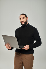 handsome and bearded arabic man in black turtleneck standing with laptop on grey background