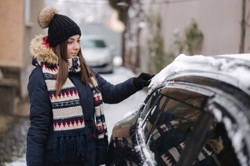 A woman is cleaning snowy window on a car with snow scraper. Pretty woman warmy dressed clean her car outdoors. Cold snowy and frosty morning. Black car