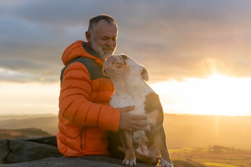 old mature man hiking in mountains with his dog, exercise and fitness for wellness, healthy lifestyle and smile. Face of a senior mature gentleman with bulldog sitting on a rock, enjoing calm day