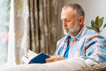 Smiling senior elderly mature man  laughing reading a book resting, sitting  on the sofa in the living room