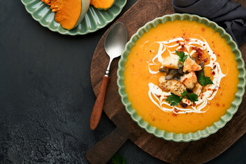 Pumpkin and carrot cream soup with herbs, seasonings and seeds in bowl on black wooden background in rustic style. Thanksgiving traditional autumn pumpkin cream soup. Top view.