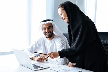 Man and woman with traditional clothes working in a business office of Dubai. Portraits of  successful entrepreneurs businessman and businesswoman in formal emirates outfits.  - obrazy, fototapety, plakaty
