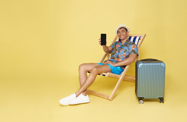 Young asian man wear summer clothes sit in deckchair showing mockup mobile cell phone app booking hotel isolated on yellow background.