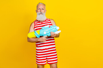Photo of serious funny old man wear striped overalls with shorts hold blue water gun on beach resort isolated over yellow color background