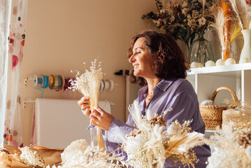 Happy mature woman working with dried flowers in floristry shop in daylight