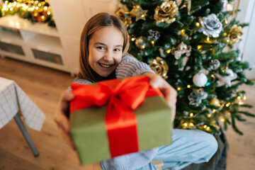Fototapeta na wymiar Closeup portrait of cheerful adorable little girl holding, giving, showing at camera wrapped with red ribbon and bow festive Christmas present gift box, sitting by glowing xmas tree, looking at camera