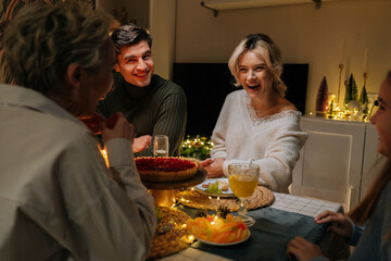 Portrait of cheerful Caucasian family celebrating Christmas, chatting at dinner holiday table in dark living room with cozy interior on xmas eve. Concept of home festive atmosphere.