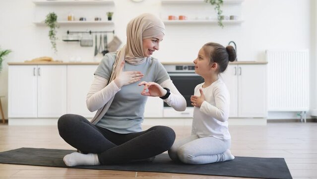Muslim female with daughter checking workout achievements on sports watch while sitting on yoga mat in kitchen. Mother and her girl taking advantages of home training using fitness tracker.
